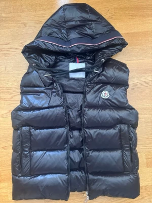 Svart dunväst med huva från Moncler - Snygg svart dunväst från Moncler med avtagbar huva och dragkedja framtill. Västen har två stora fickor och Moncler-logga på bröstet. Materialet är glansigt och quiltat, perfekt för att hålla värmen under kyliga dagar.