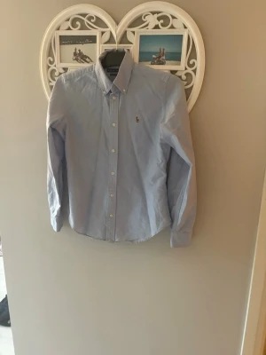 Ljusblå skjorta från Ralph Lauren - Klassisk ljusblå skjorta från Ralph Lauren med button-down krage och broderad logga på bröstet. Skjortan har långa ärmar och vita knappar framtill. Perfekt till jeans eller chinos för en clean och stilren look. Passar både xs och s