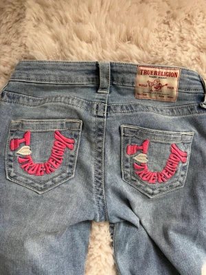 True Religion jeans - Ljusblå jeans från True Religion med ikonisk rosa brodyr på bakfickorna och detaljerade sömmar. Klassisk femficksmodell med silverfärgade nitar och bälteshällor. Snygga kontrastsömmar och patch med logga bak i midjan. Perfekta för en trendig streetwear-look. Använda ett fåtal gånger 