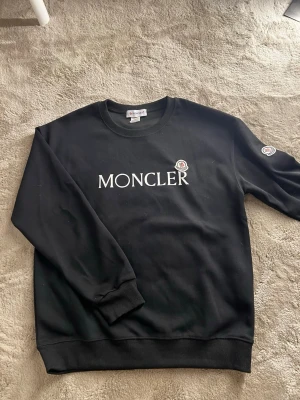 Svart Moncler sweatshirt - Snygg svart sweatshirt från Moncler med stor vit logga på bröstet och patch på ärmen. Klassisk rund halsringning och ribbade muddar vid ärmslut och nederkant. Perfekt för en clean och stilren look.