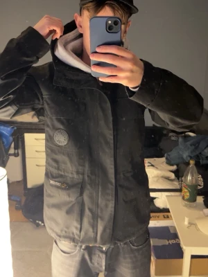 Parka jacka/fieldjacket canada goose - Cool svart vindjacka från canada goose med huva och dragkedja. Jackan har en patch på bröstet och en gul logotyp på ryggen. Materialet är slitstarkt och jackan har praktiska fickor med dragkedja. Perfekt för dig som vill ha en stilren och funktionell jacka.
