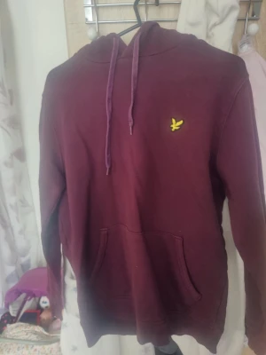 Vinröd hoodie från Lyle & Scott - Snygg vinröd hoodie från Lyle & Scott med klassisk gul logga på bröstet. Hoodien har huva med dragsko och en stor magficka framtill. Perfekt för en avslappnad och stilren look.