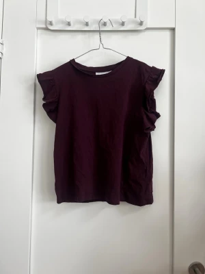 Vinröd topp med volangärmar - Supersöt vinröd topp med volangdetaljer på ärmarna. Rak passform och rund halsringning. Perfekt att styla med jeans eller kjol för en trendig look.