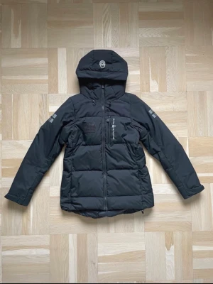 Svart dunjacka från Sail Racing - Snygg svart dunjacka från Sail Racing med huva och dragkedja framtill.                                    Ordinarie pris 3’500kr på ett ungefär!!                                                            Jackan har logga på bröstet och patch med texten 'Sail Antarctica'. Flera praktiska fickor och justerbar huva. Perfekt för kalla vinterdagar och riktigt varm. 