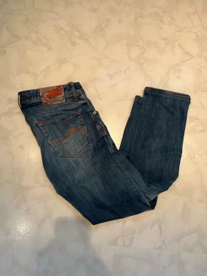  Jacob Cohen Jeans - Snygga blå jeans från Jacob Cohen med klassisk femficksmodell och orangea sömmar. Jeansen har en patch med logga bak i midjan och raka ben. Tillverkade i mjukt denimtyg som ger en avslappnad look. Perfekta för dig som gillar stilrena och tidlösa jeans.