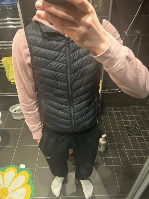 Svart dunväst med dragkedja - Snygg svart dunväst med hög krage och dragkedja framtill. Västen har ett quiltat mönster och är lätt vadderad för extra värme. Perfekt att slänga över en hoodie eller tröja för en casual look. Materialet är glansigt och ger en modern känsla. Vid väldigt noggrant sökande finns mindre fläckar som förmodligen går bort vid tvättning 