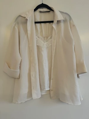 Beige skjorta med spetsdetalj - Stilren beige skjorta i lätt och luftig kvalitet med klassisk krage och knappar framtill. Under skjortan syns ett linne med spetskant vid urringningen som ger en extra detalj. Perfekt för en avslappnad men ändå chic look.