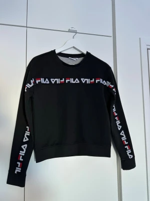 Svart sweatshirt från FILA - Säljer en svart sweatshirt från FILA med logga i vitt och rött längs bröstet och ärmarna. Tröjan har rund halsringning och ribbade muddar vid ärmslut och nederkant. Perfekt för en sportig och avslappnad stil. Tröjan har små slitningar men som man inte tänker på. 