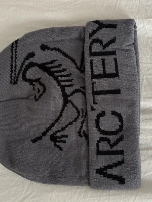 Grå mössa från Arc'teryx - Säljer en snygg grå mössa från Arc'teryx med svart logotyp och text. Mössan är stickad i mjukt material och har en uppvikt kant. Perfekt för kalla dagar och streetstyle.