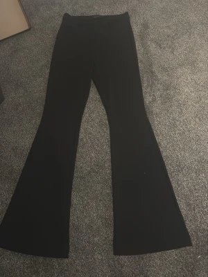 Svarta utsvängda kostymbyxor - Säljer ett par svarta kostymbyxor med utsvängda ben och hög midja. Byxorna har en klassisk look och är perfekta för dig som gillar stilrena plagg med lite edge. Materialet känns mjukt och följsamt, och de har hällor för skärp.