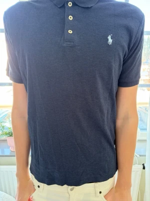 Mörkblå pikétröja från Ralph Lauren - Klassisk mörkblå pikétröja från Ralph Lauren med korta ärmar och vit broderad logga på bröstet. Tröjan har krage, knappar framtill och är tillverkad i mjuk bomull. Perfekt för en clean och stilren look.