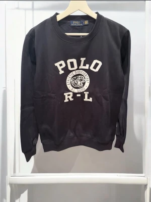  Polo Ralph Lauren tröja  - Mörkblå sweatshirt från Polo Ralph Lauren med vit text och ett tigertryck på bröstet. Klassisk rund halsringning och ribbade muddar vid ärmslut och nederkant. Perfekt för en avslappnad och sportig stil.