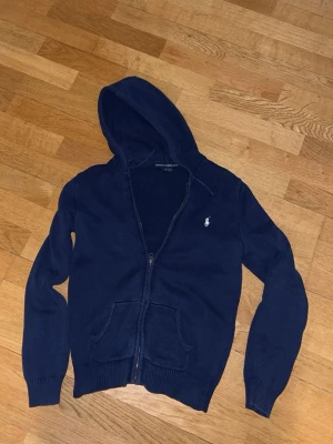 Mörkblå hoodie från Polo Ralph Lauren - Mörkblå hoodie med dragkedja från Polo Ralph Lauren. Klassisk design med huva, två fickor framtill och den ikoniska vita logon broderad på bröstet. Det är stickad. Den passar S/XS eller 170-173