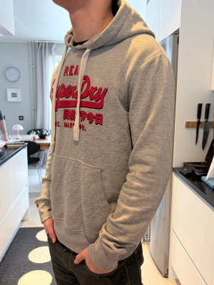Grå Superdry hoodie med röd logga - Säljer en grå hoodie från Superdry med stor röd logga och japanska tecken på bröstet. Tröjan har huva med vita snören och en stor magficka. Materialet är mjukt och bekvämt, perfekt för chill dagar. Ribbstickade muddar vid ärmslut och nederkant.