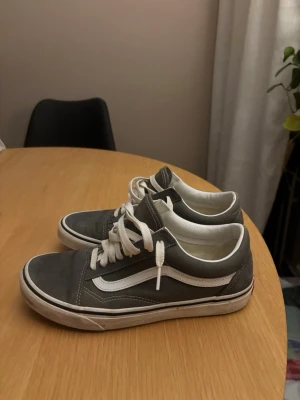 Gråa Vans Old Skool sneakers - Klassiska Vans Old Skool sneakers i grå mocka och canvas med vita detaljer och soprent sidostreck. Skorna har vita snören och en vit sula med röd Vans-logga bak. Perfekta för en avslappnad streetwear-look.
