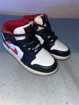 Nike Air Jordan 1 gym red Mid  - Snygga Nike Air Jordan 1 Mid sneakers i svart, vitt och rött. Skorna har klassisk design med svart läderdetaljer, vit tåbox och sidor samt röda detaljer på swoosh, foder och yttersula. Perforerad tå och ikoniska Jordan-loggor på sidan och plös.
