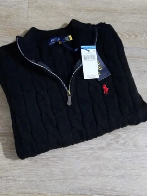 Svart kabelstickad tröja Polo Ralph Lauren - Kabelstickad svart tröja från Polo Ralph Lauren med halv dragkedja och röd broderad logga på bröstet. Tröjan har hög krage och ribbade muddar. Perfekt för dig som gillar klassisk och stilren design.