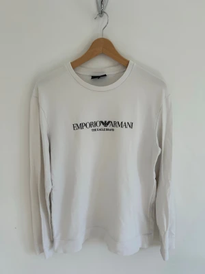 Vit sweatshirt från Emporio Armani - Vit långärmad sweatshirt från Emporio Armani med svart logotyp och text på bröstet. Klassisk rund halsringning och avslappnad passform.