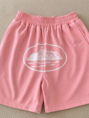 Rosa shorts med tryck och resårmidja - Säljer ett par ljusrosa shorts med elastisk midja och ett vitt tryck framtill som föreställer en byggnad eller landskap. Shortsen har en bakficka och är tillverkade i ett mjukt syntetmaterial. Perfekta för sommardagar eller chill hemma.