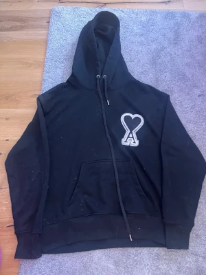 Svart hoodie från Ami Paris - Snygg svart hoodie från Ami Paris med stor huva och känguruficka. På bröstet sitter ett vitt broderat hjärta med bokstaven A. Tillverkad i mjuk bomull, perfekt för en chill och avslappnad stil.