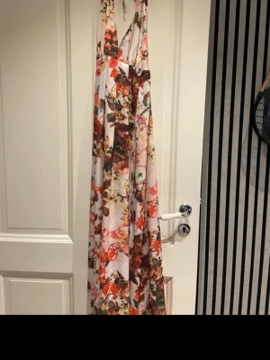 Blommig långklänning med volang - Säljer en blommig långklänning i vitt med inslag av rött, orange och grönt. Klänningen har smala axelband och ett luftigt fall med volangdetaljer. Perfekt för sommarens alla tillfällen och tillverkad i ett lätt och skönt material.