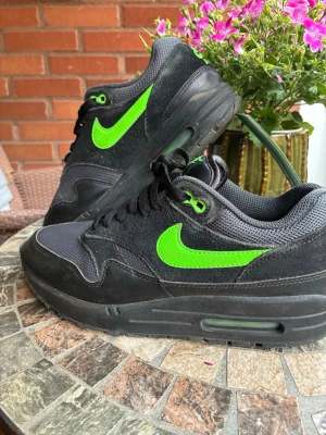 Nike Air Max 1 svart/grön sneaker - Nike Air Max 1 sneakers i svart mesh och mocka med neongröna detaljer och klassisk swoosh på sidan. Tjock sula med synlig Air-enhet och broderad Nike Air-logga bak. Skön och sportig modell som sticker ut med sin färgkombination.