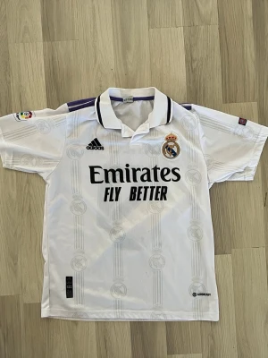 Real Madrid matchtröja Adidas vit - Säljer en vit Real Madrid matchtröja från Adidas med klubbmärke, svart Adidas-logga och 'Emirates Fly Better' tryck på bröstet. Tröjan har krage med lila och svart detalj, samt La Liga-märke på ärmen. Tillverkad i lätt och ventilerande material.