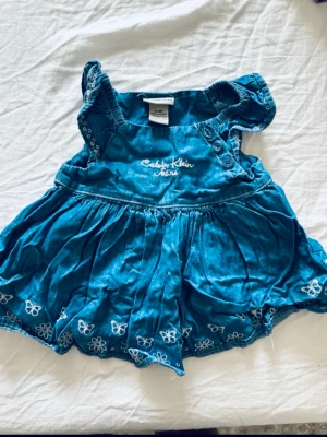 Blå babyklänning Calvin Klein Jeans - Supersöt blå babyklänning från Calvin Klein Jeans med broderade vita fjärilar och blommor längst ner. Klänningen har korta ärmar med volang och knappar på axeln för enkel påklädning. Perfekt för sommarens alla tillfällen!