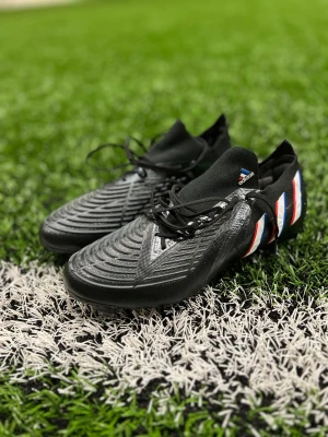 Adidas svarta fotbollsskor med snörning - Säljer ett par svarta Adidas fotbollsskor med snörning och stickad strumpkrage. Skorna har mönstrad ovandel för bättre bollkontroll och klassiska Adidas-ränder i vitt, rött och blått på hälen. Perfekta för konstgräs och matcher.