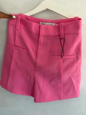 Rosa shorts från Zara med fickor - Säljer ett par snygga rosa shorts från Zara med hög midja och bälteshällor. Shortsen har två dekorativa fickor fram och är gjorda i ett mjukt material som känns bekvämt. Perfekta för varma dagar och ger en färgstark touch till din outfit.