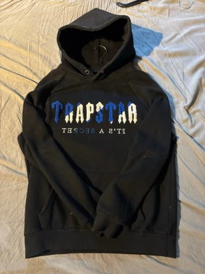 Trapstar svart hoodie med brodyr - Säljer en svart hoodie från Trapstar med stor broderad logga i blått och vitt på bröstet samt texten 'IT'S A SECRET' under. Perfekt för dig som gillar streetwear och vill sticka ut. Tvättar tröjan innan jag skickar iväg den ☺️