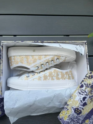 Beige och vita Dior sneakers - Säljer ett par snygga låga sneakers från Dior i beige och vitt med det klassiska Dior-mönstret. Skorna har vita snören, gummisula och detaljer med Christian Dior-logga på sidan och tungan. Perfekta för dig som vill ha en exklusiv touch till din outfit.