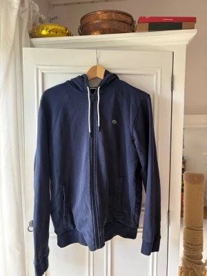 Mörkblå hoodie från Lacoste - Mörkblå hoodie från Lacoste med dragkedja framtill, vita dragsnören och klassisk krokodillogga på bröstet. Hoodien har fickor på sidorna och ribbade muddar vid ärmslut och nederkant. .