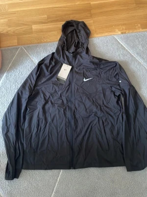 Nike Miller svart  - Snygg svart vindjacka från Nike med hel dragkedja och huva. Jackan har en minimalistisk design med Nike-logga på bröstet och är tillverkad i lätt, vindtätt material. Perfekt för blåsiga dagar och sportiga outfits.