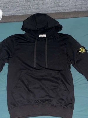 Svart hoodie från Stone Island - Säljer en svart hoodie från Stone Island med klassisk patch på vänster ärm. Hoodien har huva med dragsko och en stor magficka framtill. Tillverkad i mjukt bomullsmaterial som är skönt att bära. Perfekt för dig som gillar streetwear och vill ha en stilren look.