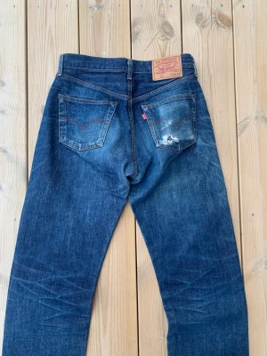 Selvedge Levi's 502 Big E Distressed - Säljer ett par klassiska blå Levi's 502 jeans med raka ben och normal midja. Jeansen har slitningar och en mindre reva på ena bakfickan, samt den ikoniska röda Levi's-lappen. Tillverkade i kraftig denim med snygg tvätt och kontrastsömmar.