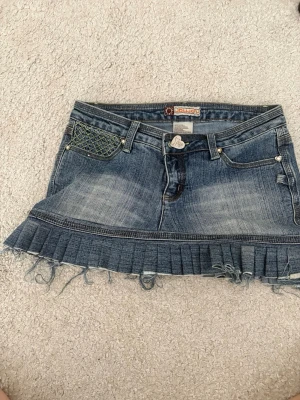 Blå jeanskjol med volang och detaljer - Säljer en blå jeanskjol från Moujonjon med volangkant och slitna detaljer. Kjolen har dekorativa fickor med broderi och nitar både fram och bak samt ett hjärtformat knapp. Perfekt för dig som gillar unik denimstil!