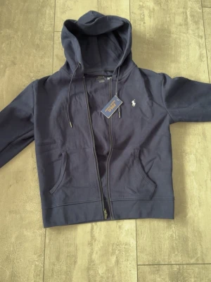 Mörkblå hoodie från Polo Ralph Lauren - Snygg mörkblå hoodie från Polo Ralph Lauren med dragkedja, två fickor framtill och klassisk huva med dragsko. Ikoniska vita loggan broderad på bröstet. Tillverkad i mjuk bomull för skön känsla och avslappnad stil.