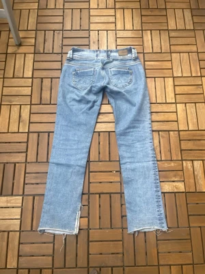 Ljusblå raka jeans med råa kanter - Säljer ett par ljusblå jeans med raka ben och råa, fransiga benslut. Jeansen har klassiska bakfickor och en avslappnad passform. Perfekta för en trendig och avslappnad stil. Materialet är denim i bomull. Passar någon som är 154