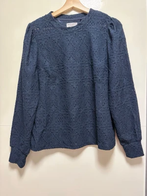 Mörkblå spetsblus från Holly & Whyte - Mörkblå blus från Holly & Whyte med långärmad design och vackert spetsmönster över hela plagget. Blusen har rund halsringning och lätt puffade axlar för en trendig siluett. Perfekt för dig som gillar detaljer och en stilren look.