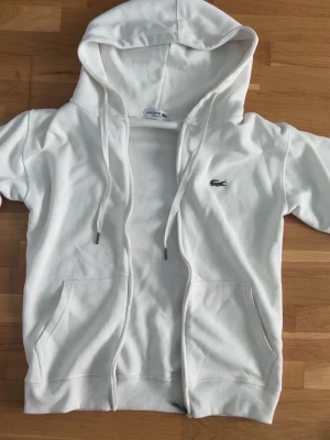 Vit hoodie från Lacoste med dragkedja - Snygg vit hoodie från Lacoste med dragkedja framtill och klassisk krokodillogga på bröstet. Regular fit-modell med två fickor och justerbar huva med snören. Tillverkad i mjukt bomullsmaterial som känns skönt mot huden.