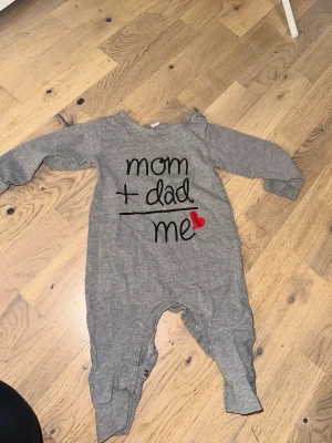 Grå babybody med text och hjärta - Grå långärmad babybody med trycket 'mom + dad = me' och ett rött hjärta. Bodyn har tryckknappar vid axeln och i grenen för enkel påklädning. Mjuk och bekväm bomullskänsla, perfekt för små barn.