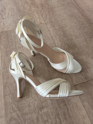 Sandalett i satin, kryssrem och rosett bak st 36 - Sandalett i satin med öppen tå och smala remmar som korsas framtill. Färg Ivory Skorna har en elegant rosett bak på hälen och justerbart spänne vid ankeln. Perfekta för dig som vill ha en chic och feminin look.