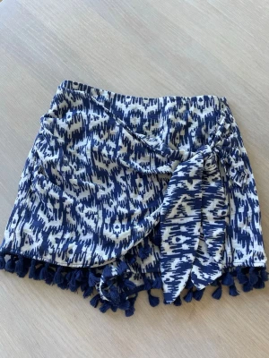Blåvita skorts med mönster och tofsar - Supersnygga blåvita skorts med grafiskt mönster och omlottkänsla. Midjan är elastisk och nederkanten är dekorerad med mörkblå tofsar för en extra cool touch. Perfekta för sommaren och riktigt bekväma. Materialet känns lätt och luftigt.