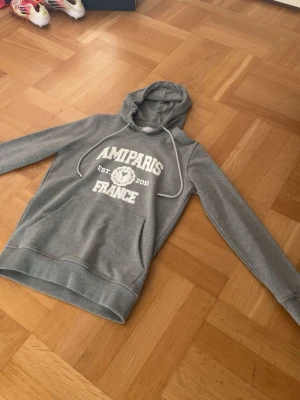 Grå hoodie AMIPARIS France - Snygg grå hoodie från AMIPARIS med vit text och tryck framtill. Klassisk känguruficka, huva med vita snören och mjukt material. Perfekt för en chill och avslappnad stil.