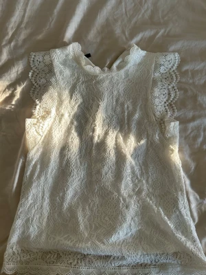 Vit spetsblus från Pieces XS - Söt vit blus från Pieces i storlek XS, helt täckt av spets med fina virkade detaljer vid ärmslut och hals. Blusen är ärmlös och har knapp i nacken för enkel påklädning. Perfekt för en fräsch och romantisk look. Köpt på Vero Moda