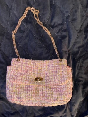 Pastellfärgad tweed axelväska - Axelväska i pastellfärgad tweed med inslag av rosa, lila och gult. Väskan har en guldfärgad kedjerem och ett vridlås i metall på framsidan. Klassisk rektangulär form och mjuk struktur, perfekt för dig som gillar en chic och trendig look. Den har lite svarta fläckar på insidan men inga  defekter på utsidan 
