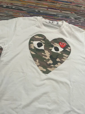 Vit t-shirt med camo-hjärta från CDG  Play - Vit t-shirt med stort hjärta i camouflage-mönster och ögon på bröstet. Liten röd hjärtdetalj med öga inuti det stora hjärtat. Från märket  Comme des Garçons play Klassisk rund hals och korta ärmar. 