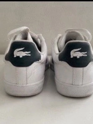 Vita Lacoste sneakers med gröna detaljer - Snygga vita sneakers från Lacoste med gröna detaljer och den klassiska krokodilloggan på sidan och hälen. Skorna har lågt skaft, vita snören och är tillverkade i en mix av läder och textil. Perfekta för en clean och sportig look.