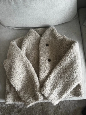 Stilren teddyjacka - Mysig beige teddyjacka med två stora bruna knappar framtill. Jackan har bred krage och är gjord i fluffigt, mjukt teddy-material som ger en avslappnad och trendig look. Perfekt för kyliga dagar när du vill vara både varm och snygg.